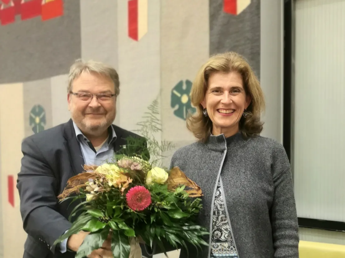 Ein Mann und eine Frau stehen vor einem Wandteppich, der Symbole und Farben der Landeshauptstadt Hannover zeigt (gr&uuml;ne Kleebl&auml;tter und rot-wei&szlig;e Wimpel). Der Mann &uuml;berreicht der Frau einen Blumenstrau&szlig;. Beide l&auml;cheln in die Kamera.